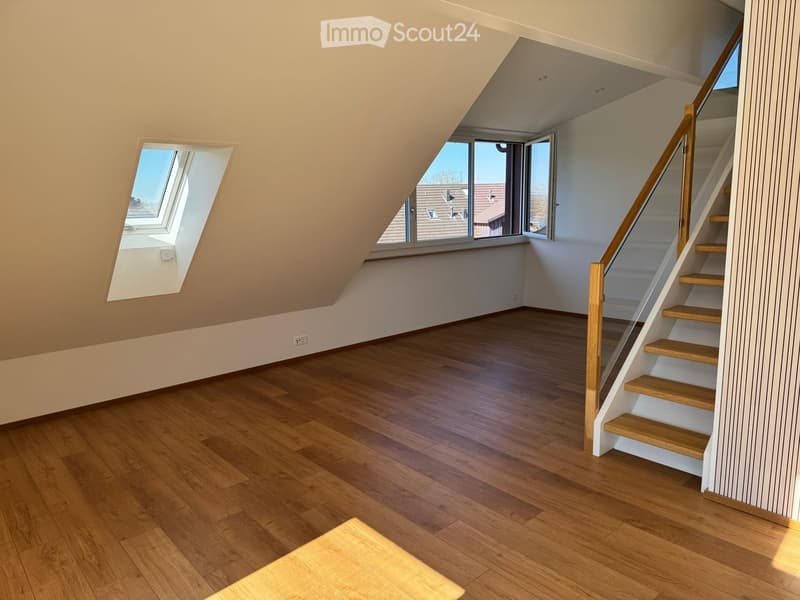 Empty attic room with wooden flooring, slanted ceilings, and a staircase leading upwards. Features large windows and skylights allowing natural light to enter.