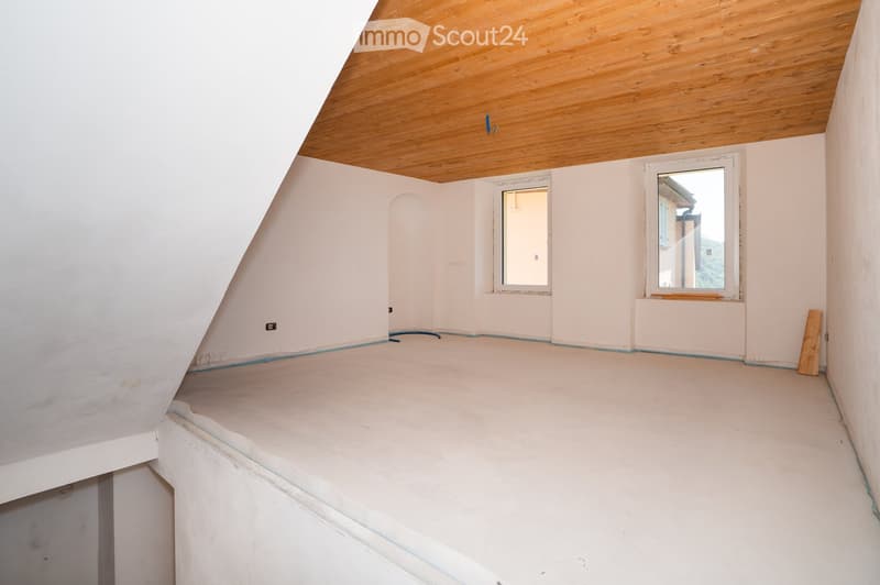 Empty attic room with wooden slanted ceiling, white walls, and two windows. The floor is unfinished concrete, and there are no furnishings or decorations.