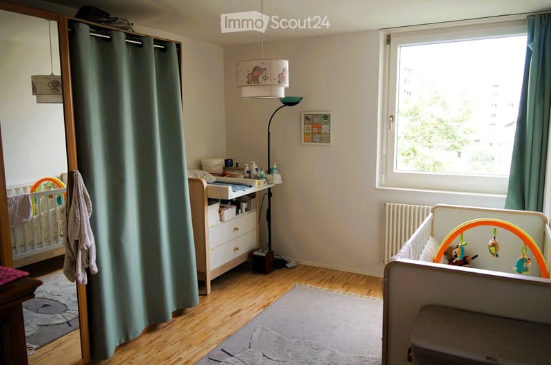 Brightly lit nursery with wooden flooring, two cribs, a changing table, a toy arch, and decorative curtains.