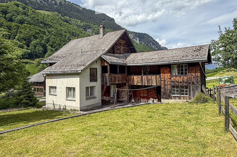 Mountain house with wooden exterior, a chimney, glass windows, and located in a valley with mountains in the background.