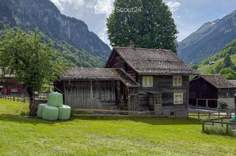 Wooden alpine house, stone chimney, wooden fence, green hay bales, mountains, grass field