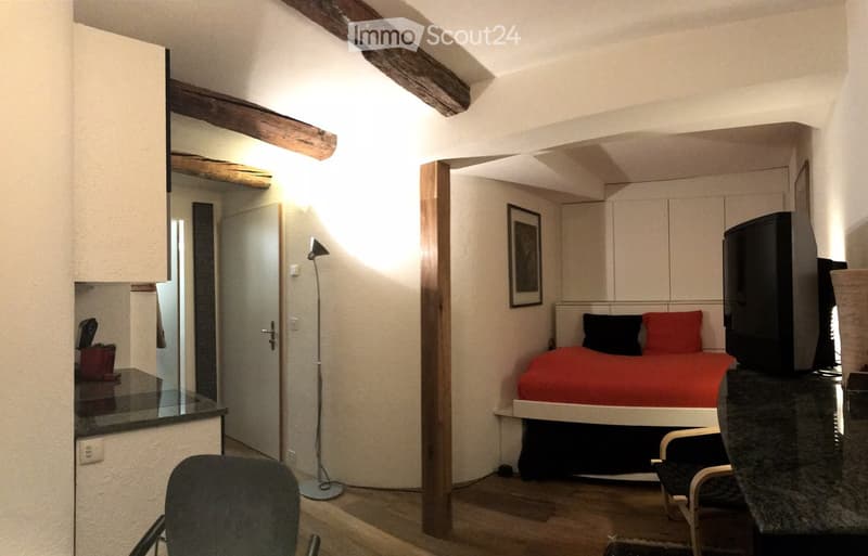 Cozy bedroom with a bed, red bedding, a chair, a table, a mirror, a floor lamp, and a television. The room features exposed wooden beams and a small seating area.