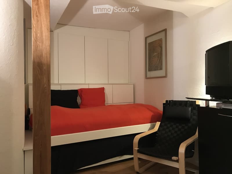 Modern bedroom with a red duvet, black pillows, a sleek white bed frame, a black and white chair, a television on a stand, and built-in white cabinets.
