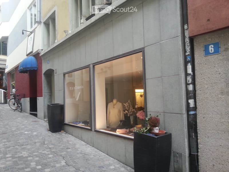 Storefront with large glass windows displaying clothing and accessories, including a mannequin, flowers, and decorative items. The building has a modern facade with grey stonework and a blue awning on the adjacent building.