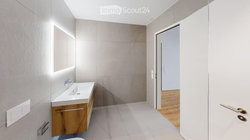 Modern bathroom with a white sink, wooden cabinet, and large mirror illuminated by LED lighting. The space features light grey tiles and a clean, minimalist design.