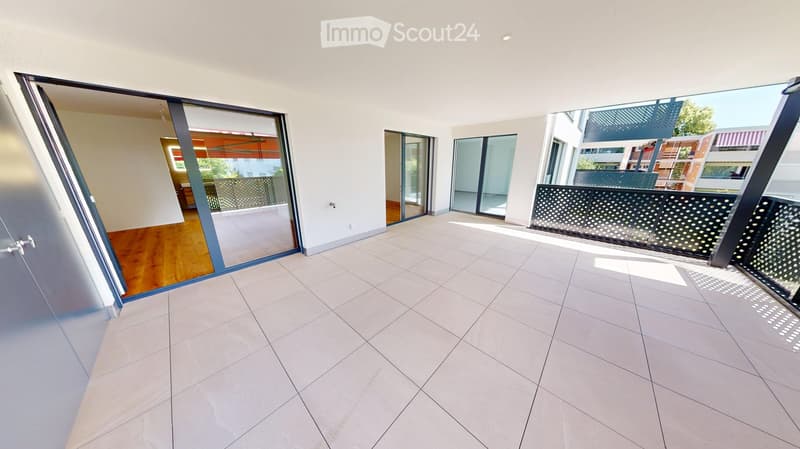 Modern balcony with large glass sliding doors, tiled floor, and metal balustrade with perforated design. The space is open and bright, with views of neighboring buildings.