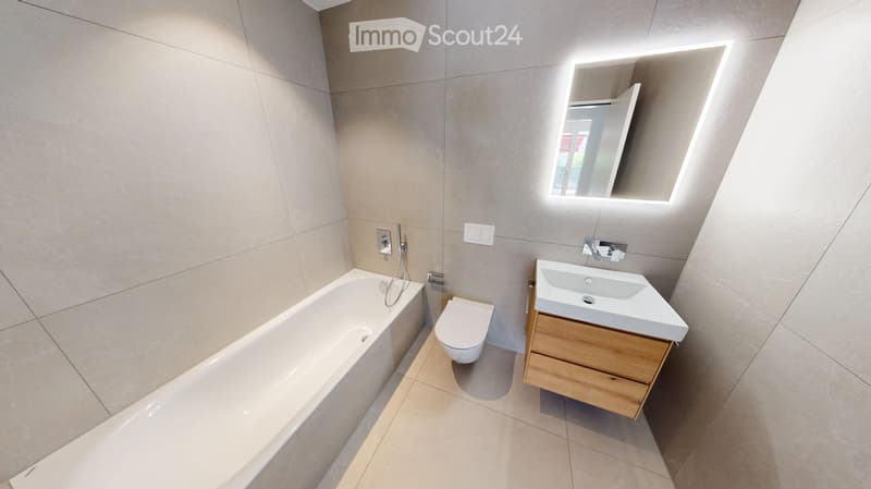 Modern bathroom with a bathtub, toilet, sink, and a wooden cabinet. Features a large mirror with built-in lighting and sleek, contemporary design.