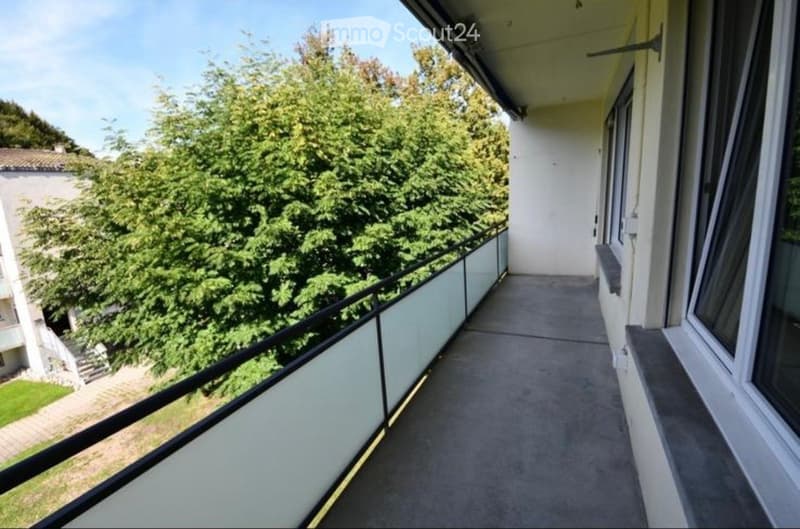 Balcony with a view of green trees and a neighboring building, featuring a metal railing and a concrete floor.