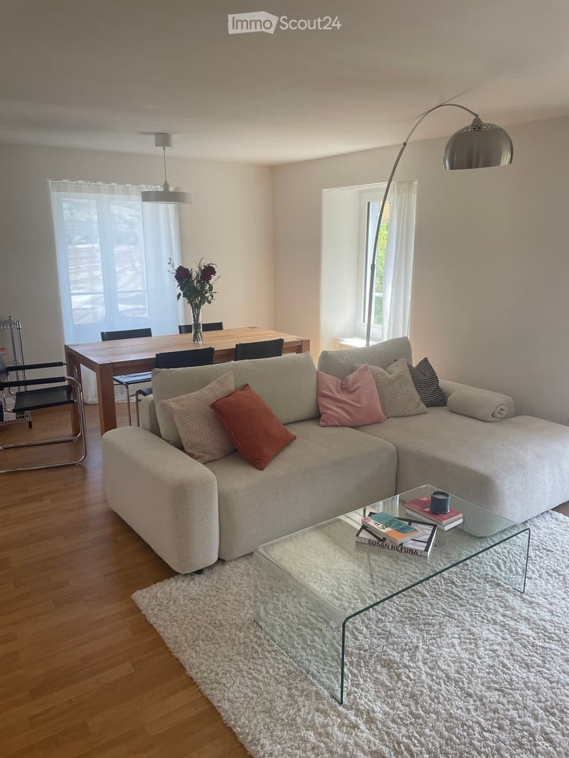 Modern living room with a grey sectional sofa, glass coffee table, dining area with wooden table and chairs, and a vase with flowers on the table.