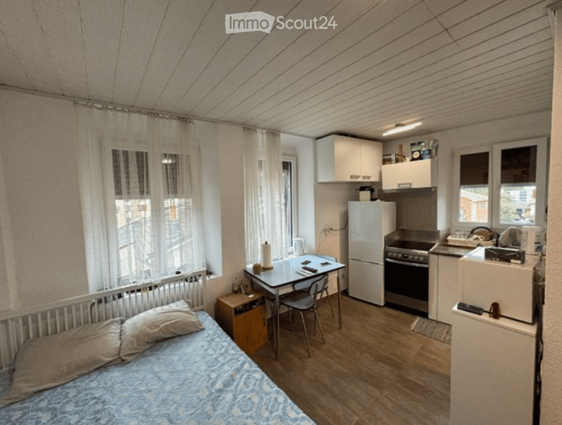 A compact studio apartment with a bed, small table, chairs, and a kitchenette area featuring a refrigerator, oven, microwave, and cabinets.