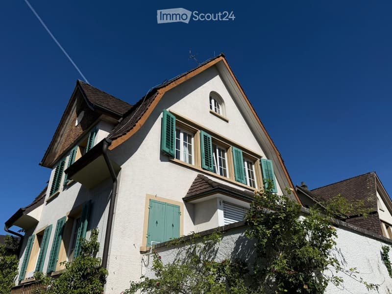 Two-story house with white walls, green shutters, and a brown roof under a clear blue sky.