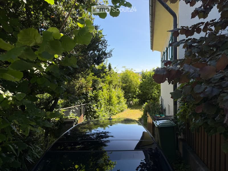 A narrow garden path lined with trees and a parked car, with a building on the right side.