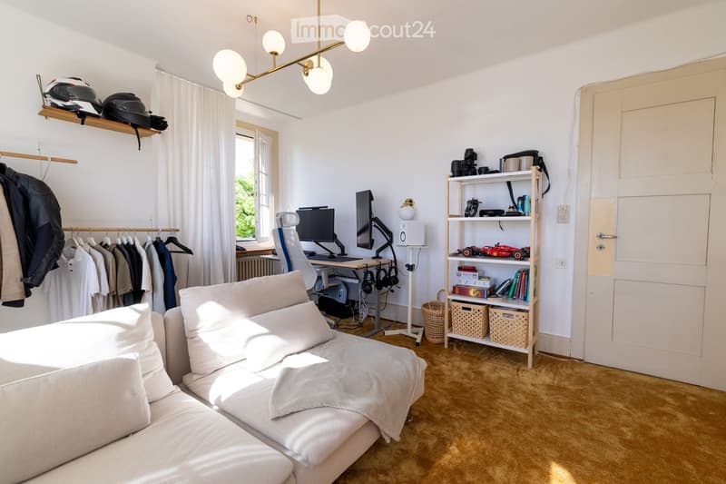 A well-lit, multi-functional room with a sofa, desk, and shelves. Features include a hanging light fixture, a window with curtains, and a clothing rack with jackets.
