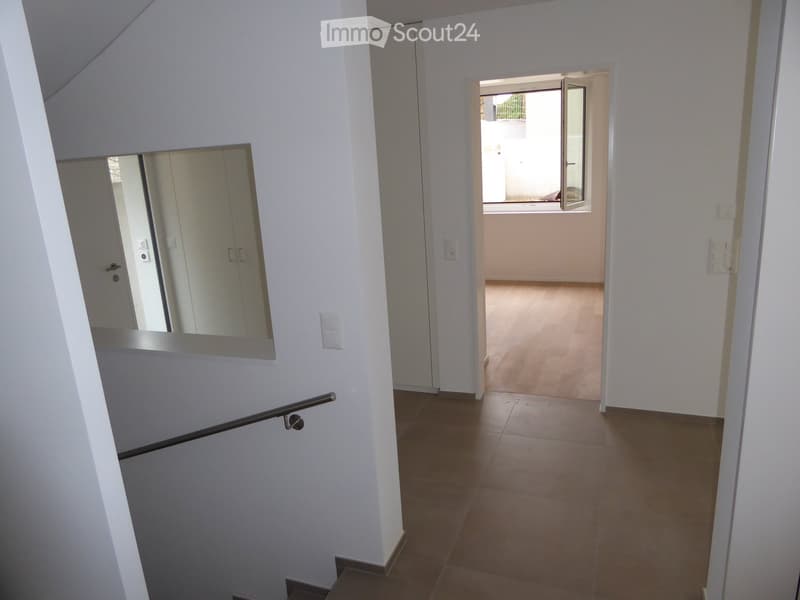Corridor / Hallway. A modern, clean hallway with white walls, tiled floors, and a staircase leading upwards. A large mirror is mounted on the wall, and an open doorway leads to another room with light-colored flooring and a visible window.