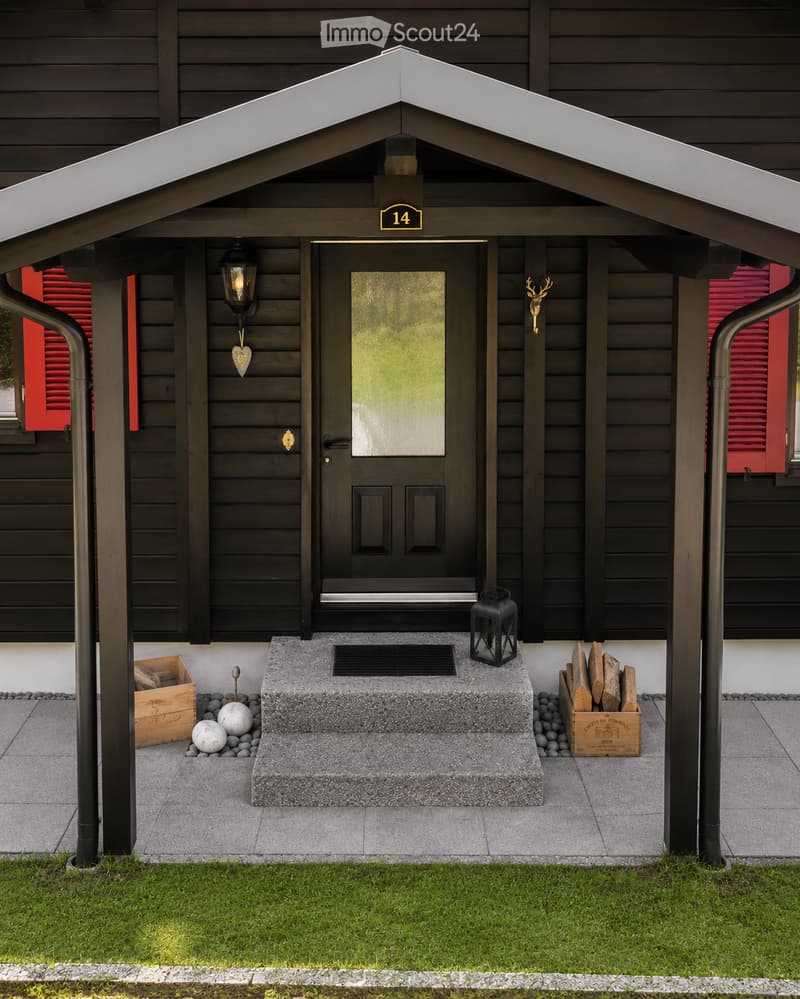 Front entrance of a house with a covered porch, black door, lantern-style light fixture, decorative deer head, and stacked firewood crates.