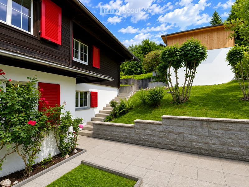 Two-story house with red shutters, white walls, and a stone pathway leading to a raised garden area with green grass and shrubs.