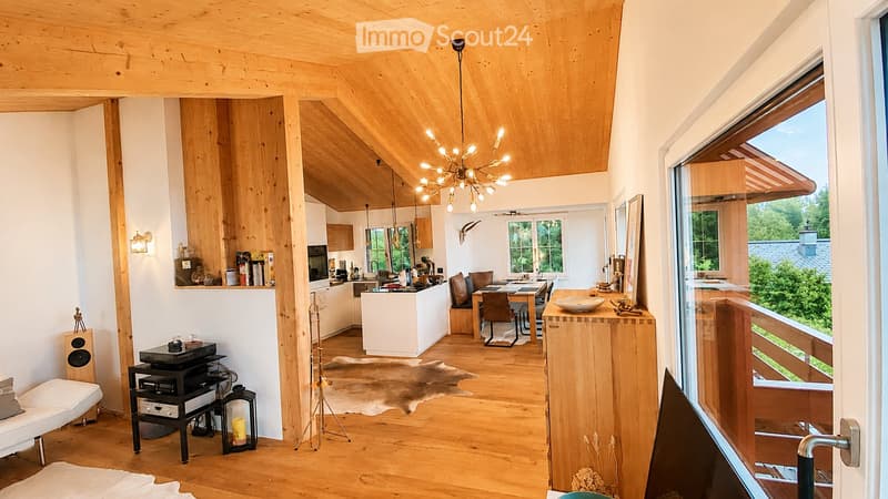 Open-plan living and dining area with wooden floors, exposed wooden beams, large windows offering a view, a white sofa, coffee table, kitchen island, dining table with chairs, chandelier, speaker, and decorative items.