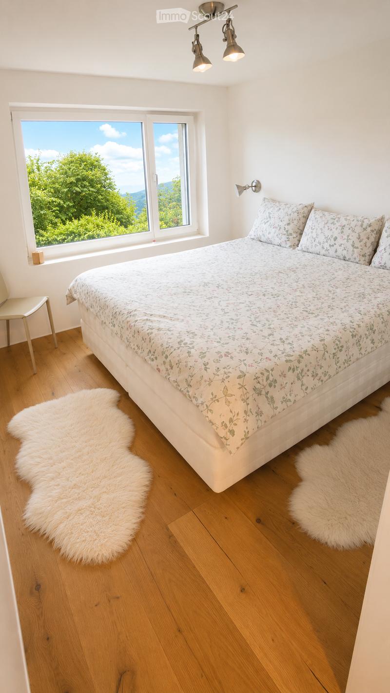 Bright bedroom with wooden flooring, large window offering a scenic view, a neatly made bed with floral bedding, a white fur rug, and a small chair near the window.