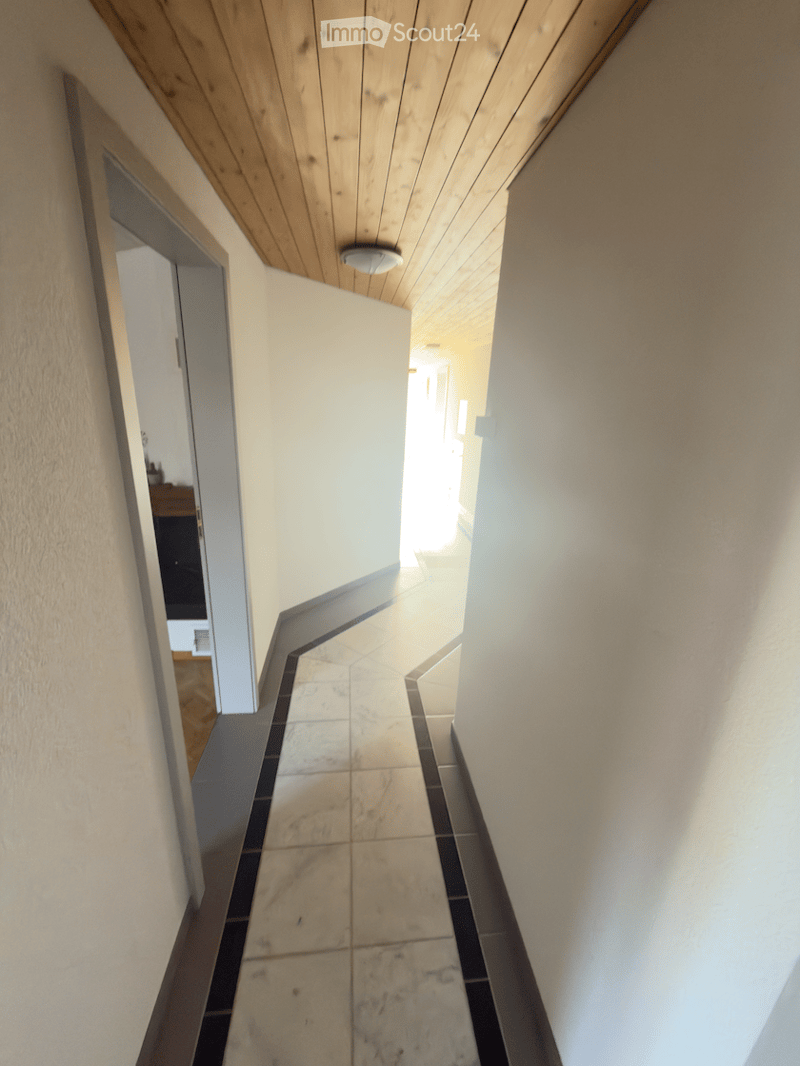 A narrow hallway with tiled flooring and wooden ceiling, leading to an open area with natural light.