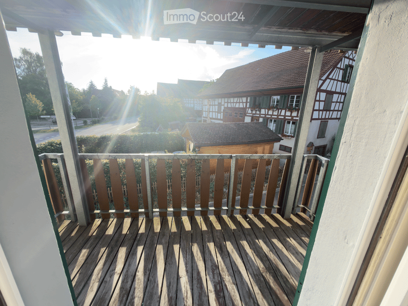 Wooden balcony with railing, overlooking a street with traditional houses and greenery.