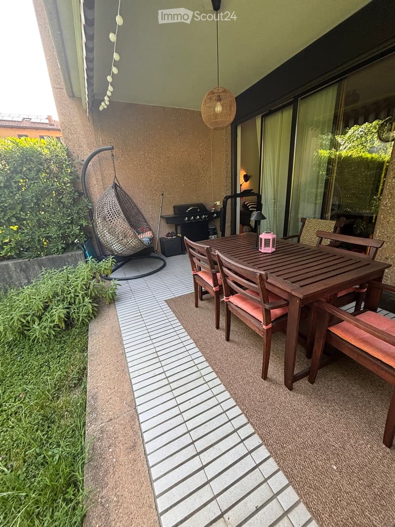 Balcony with tiled floor, wooden table and chairs, wicker hanging chair, and grill.