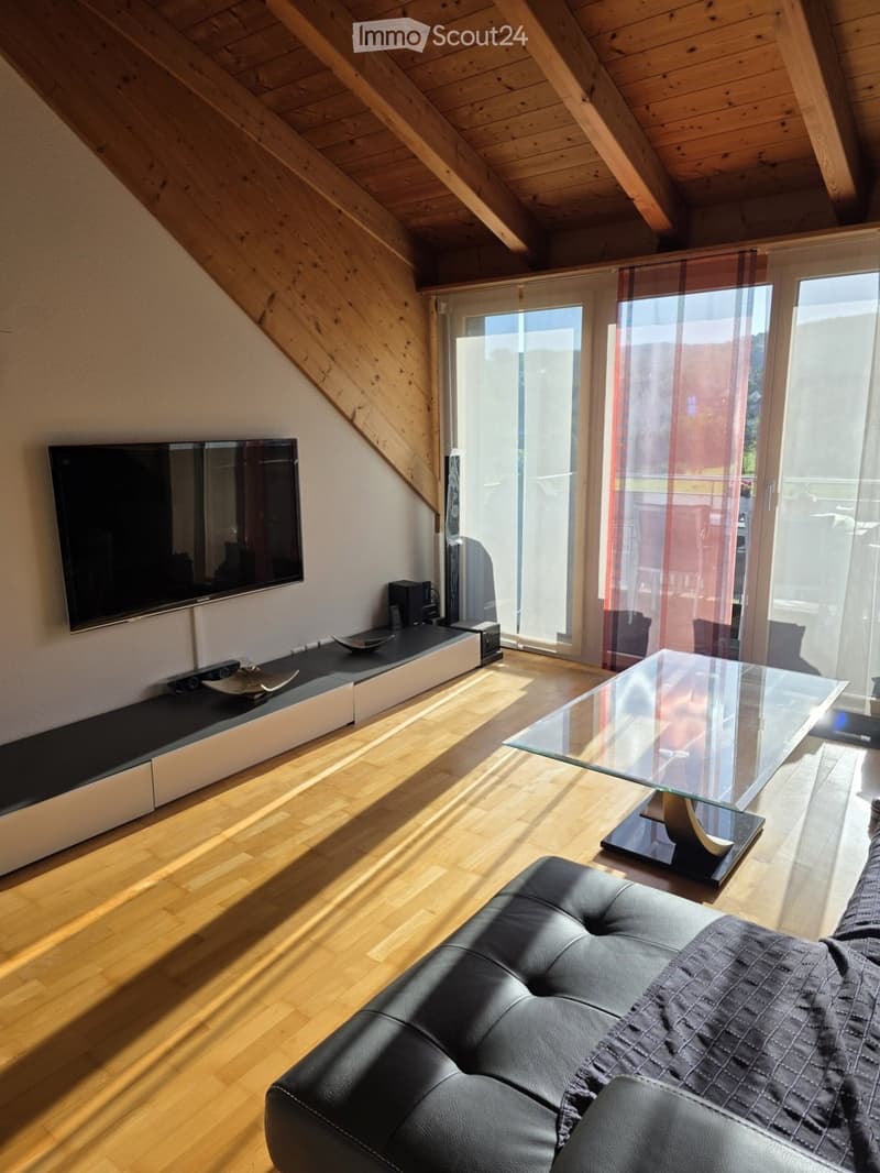 Modern living room with wooden flooring, a black leather sofa, a glass coffee table, and a flat-screen TV on a sleek entertainment unit. Large windows with sheer curtains allow natural light to fill the space.