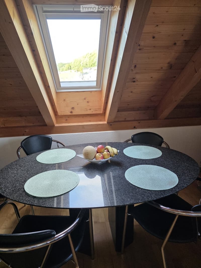 A round dining table with four chairs, placemats, and a bowl of fruit on the table. The room has wooden walls and a large window letting in natural light.