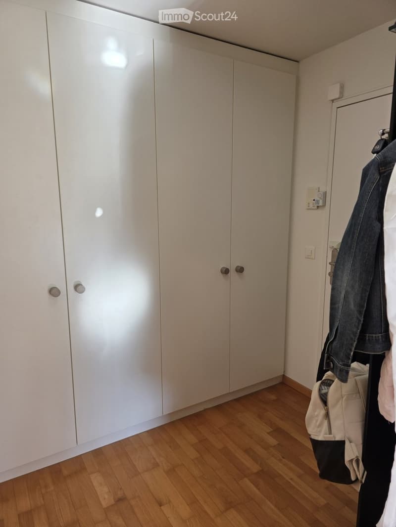 Empty room with light-colored wooden flooring and white built-in wardrobes. A coat and bag are hanging on a rack near the door.