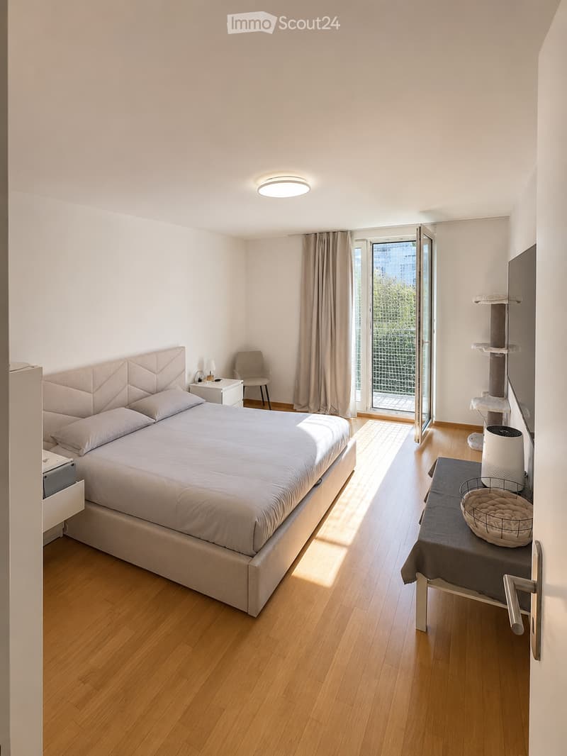 Modern bedroom with a large bed, nightstand, chair, cat tree, and a small table with a cat bed on it. The room features wooden flooring, large windows with curtains, and a sleek, contemporary design.