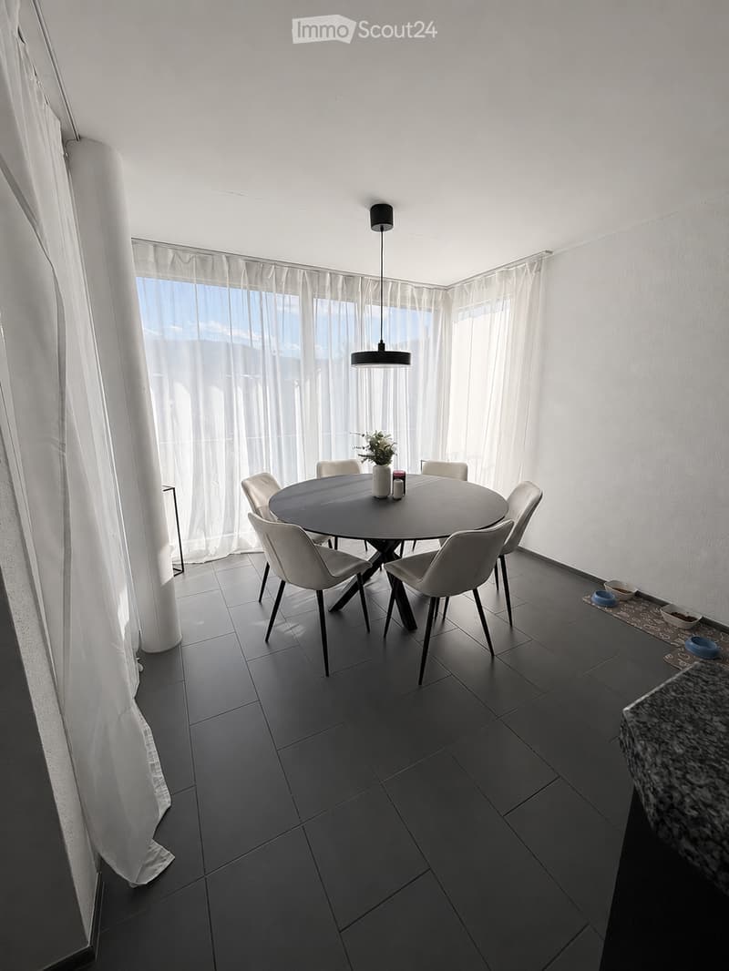 Modern dining area with a round black dining table surrounded by six white chairs, a pendant light fixture, and a small side table with a plant and candles.