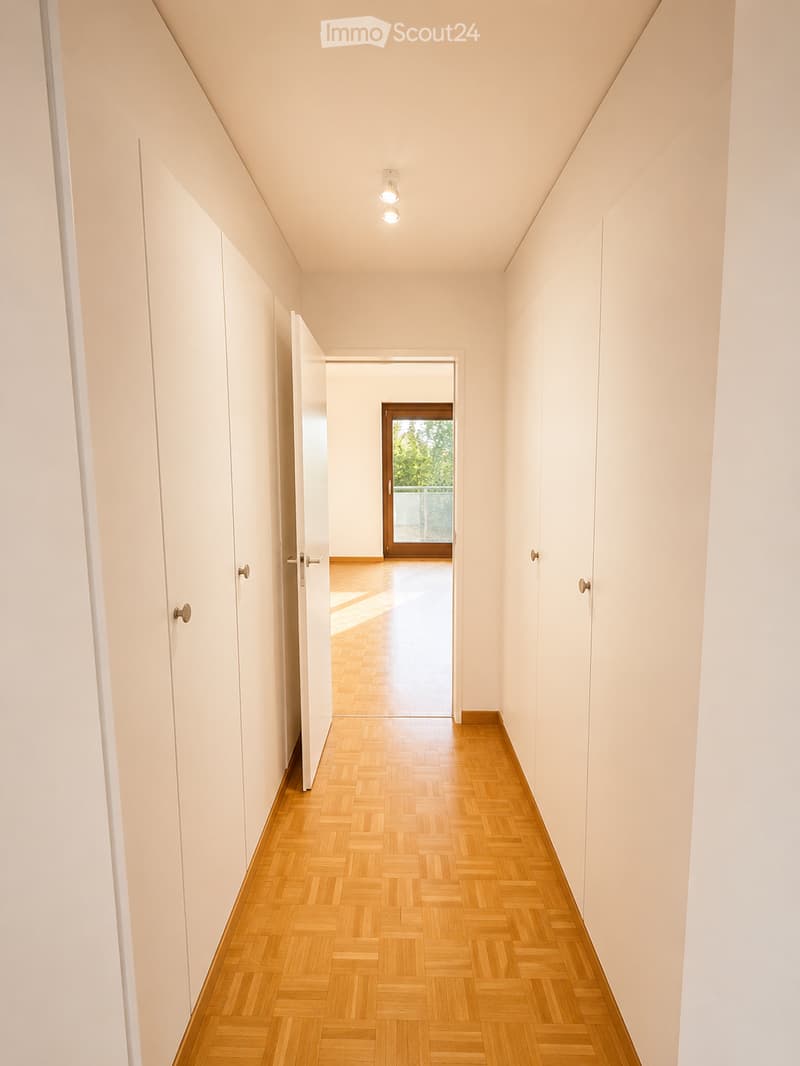 A long, narrow hallway with light-colored walls, wooden herringbone flooring, and recessed ceiling lights. The hallway features several closed white doors on both sides and leads to an open doorway at the end.