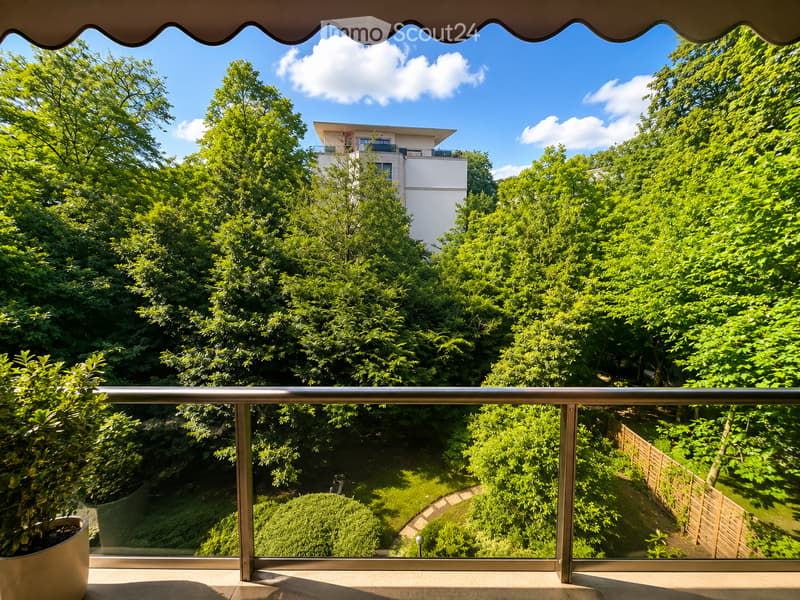 Balcony with metal railing, overlooking a lush green garden and a modern white building in the background.