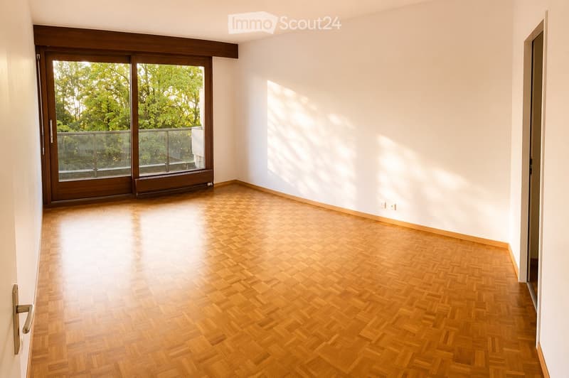 Empty room with wooden herringbone flooring, white walls, large sliding glass door leading to a balcony, and natural light streaming in.