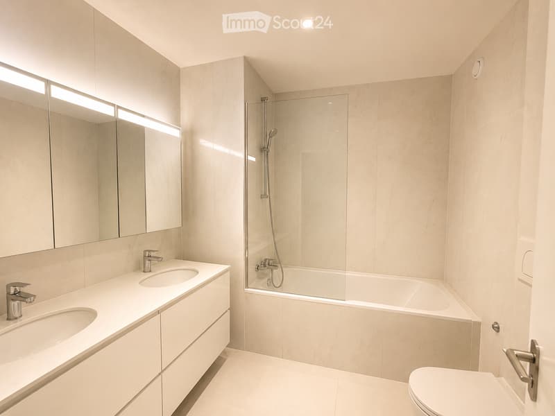 Modern bathroom with two sinks, a bathtub, a toilet, and a large mirror with integrated lighting. The space features sleek, contemporary design with light-colored tiles and a glass-enclosed shower area.