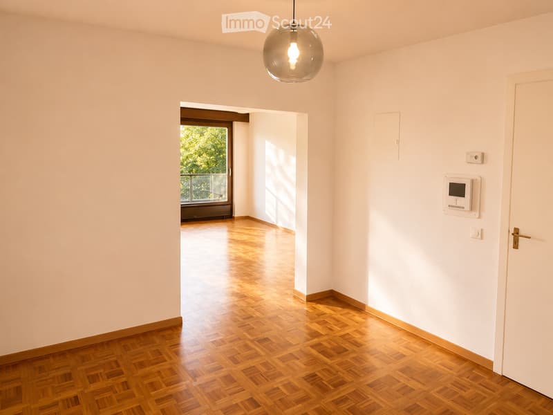 Empty room with light-colored walls, wooden parquet flooring, and a pendant light fixture. A doorway leads to a balcony area with a view of greenery.