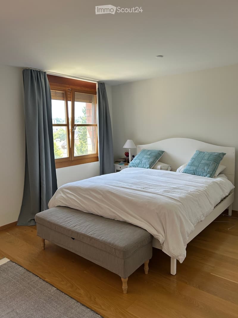 Cozy bedroom with a large bed, bench at the foot, nightstand with lamp, and grey curtains on the window.