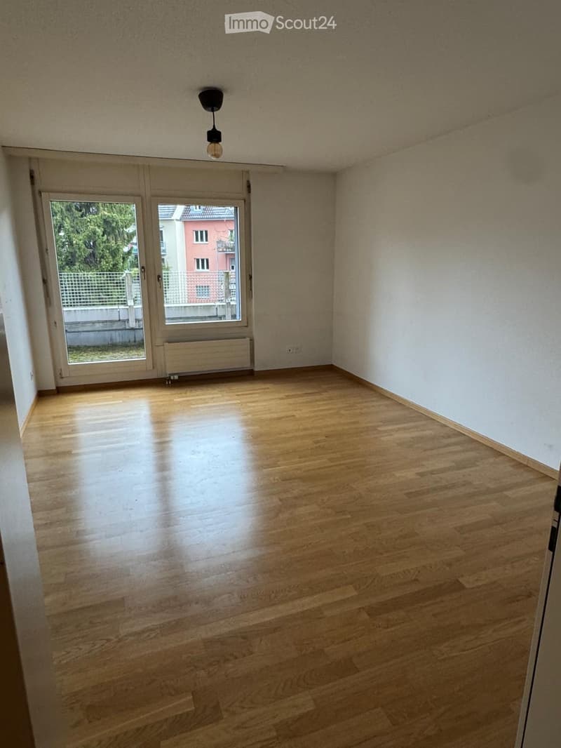 Empty room with wooden flooring, white walls, and a large window with a view of the outside. There is a radiator below the window and a ceiling light fixture.