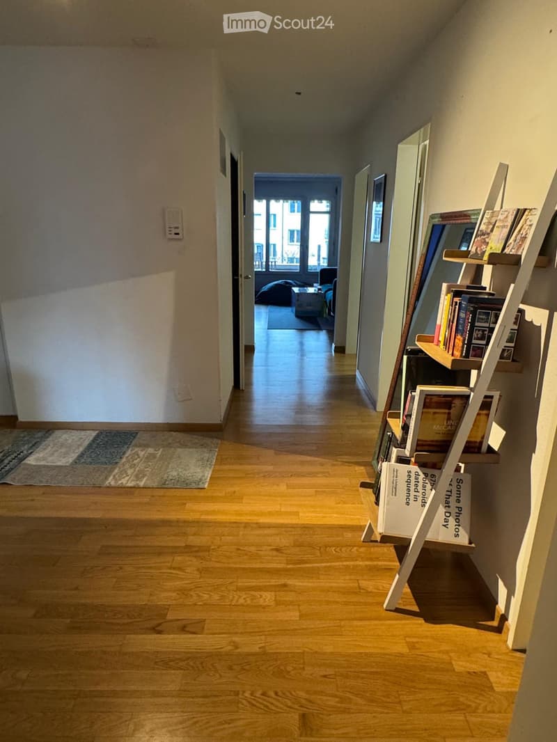 A well-lit hallway with wooden flooring, a small rug on the left, and a leaning bookshelf with books and framed pictures on the right. The hallway leads to a room with a sofa and coffee table visible in the background.