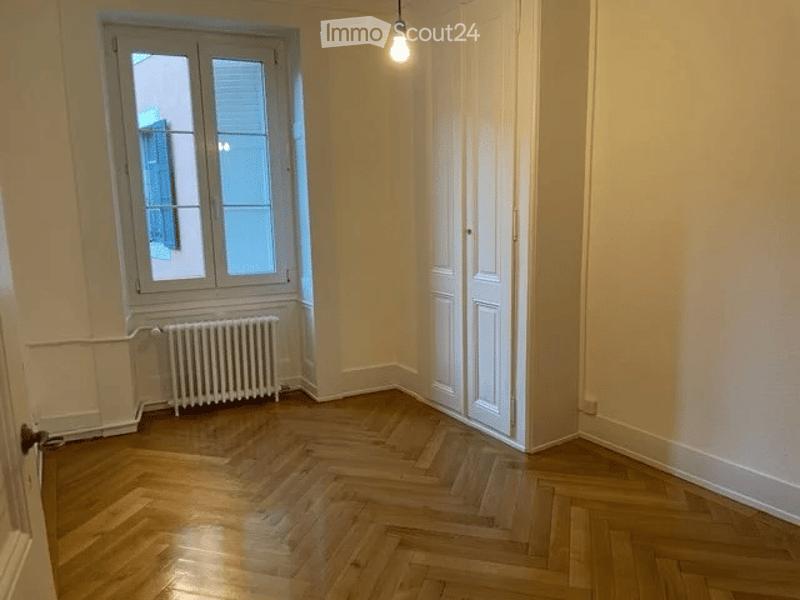 Empty room with wooden herringbone flooring, white walls, a single window with shutters, a radiator, and a closed white door.