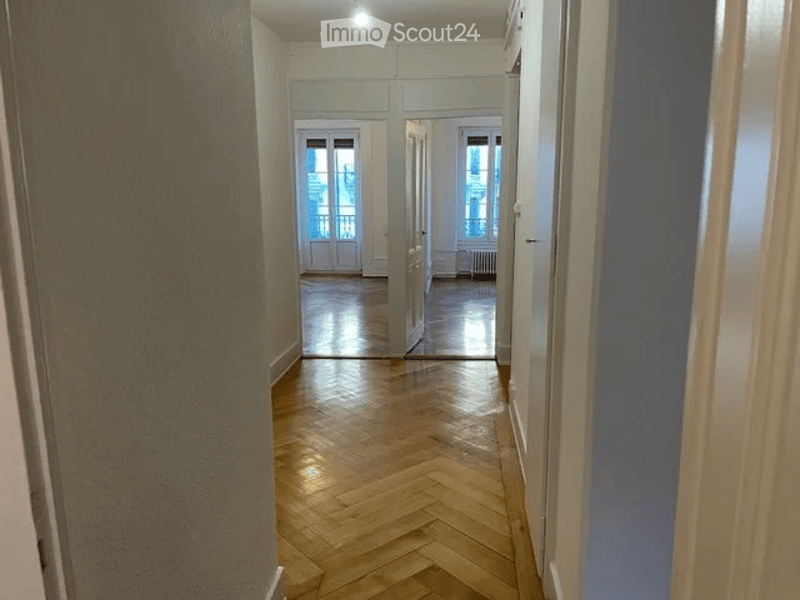 Empty hallway with wooden herringbone flooring, white walls, and doors leading to rooms.