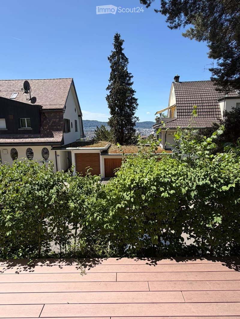 A sunny outdoor patio with a tiled surface, surrounded by lush green bushes and trees, with residential houses and a clear blue sky in the background.