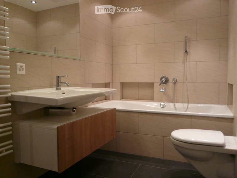 Modern bathroom with beige tiles, a double sink, a bathtub, a toilet, a mirror, and a radiator.