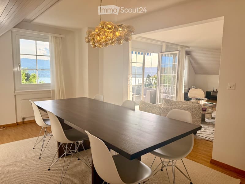 Bright dining area with a large wooden table, white chairs, and a modern chandelier. Adjacent living area features a patterned sofa, coffee table, and scenic mountain view through large windows and sliding doors.