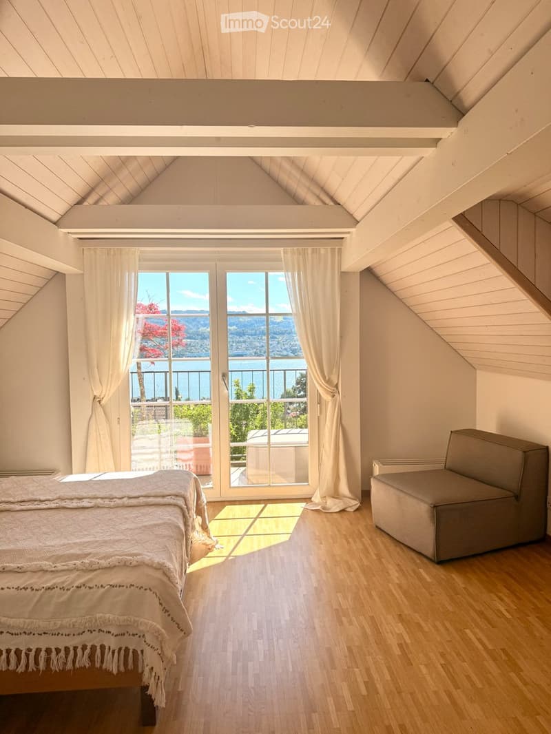Bright bedroom with wooden flooring, large window offering a scenic view, bed with white bedding, and a small chair.