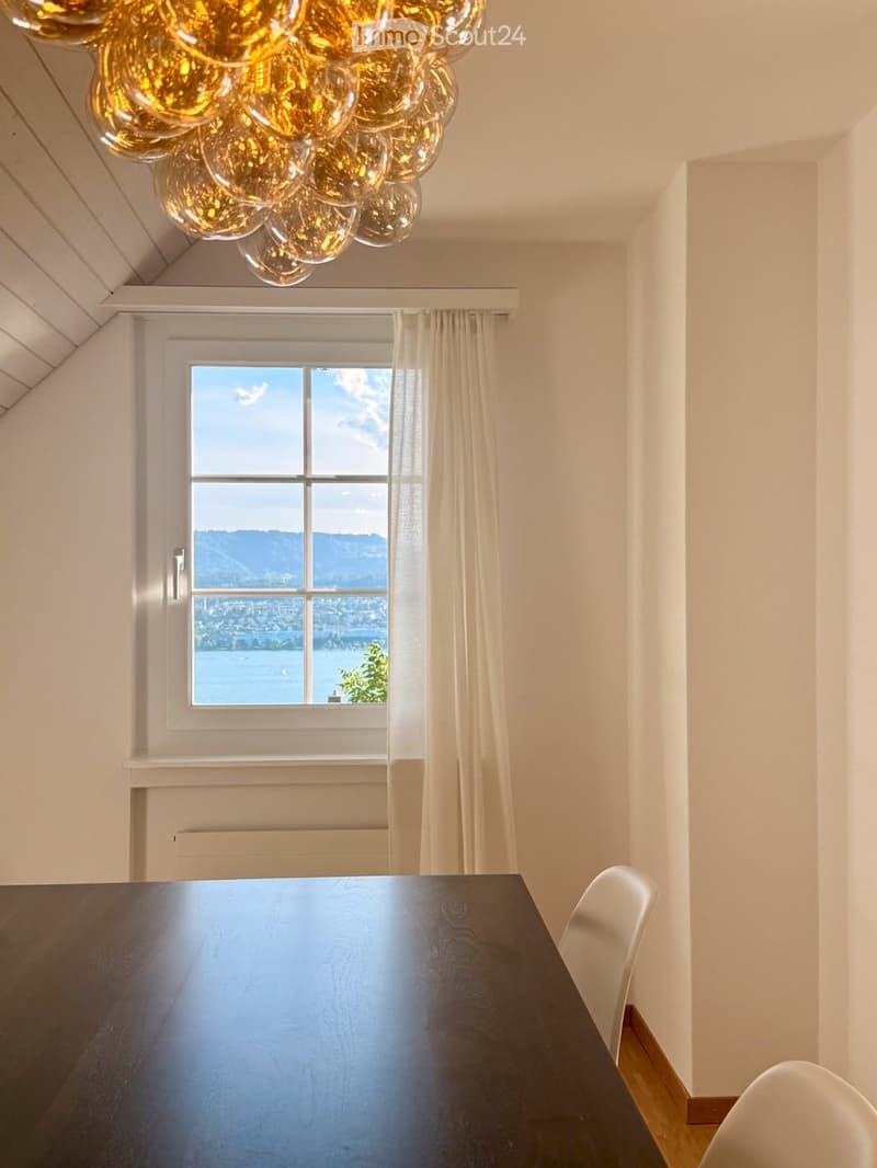 Bright dining area with a dark wooden table, white chairs, large window with a scenic view of a lake and mountains, and a modern chandelier.