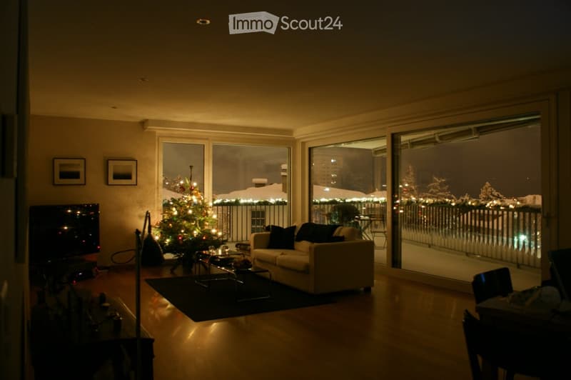 Cozy living room with a sofa, coffee table, television, and a decorated Christmas tree. Large windows offer a view of a snowy outdoor scene.