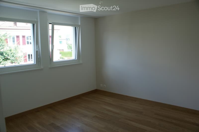 Empty room with wooden flooring, white walls, and two large windows with white frames, allowing natural light to enter.