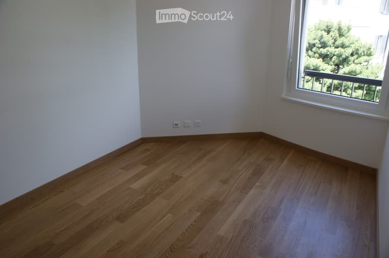 Empty room with light wooden flooring and white walls, featuring a large window with a view of greenery and a balcony outside.
