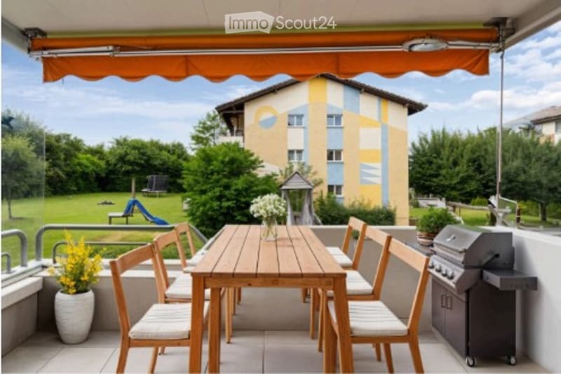 Balcony with wooden table, chairs, potted plants, and a barbecue grill. The background shows a green park area with trees and a playground.
