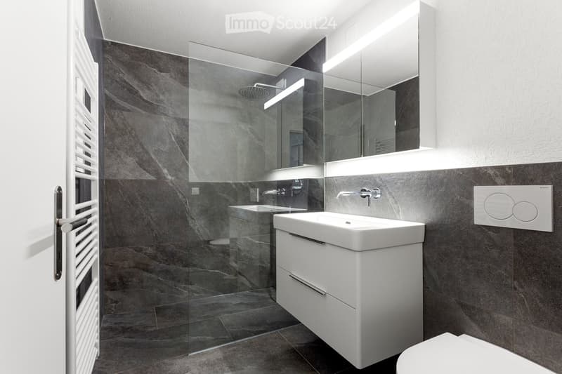 Modern bathroom with dark grey marble tiles, white sink with integrated storage, large mirror with built-in lighting, glass-enclosed shower, and a radiator on the left side.